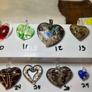 May include: A collection of heart-shaped glass pendants in various colors and designs. Some pendants have metallic accents, while others feature swirls, stripes, and floral patterns. Each pendant has a small loop for hanging and is numbered.