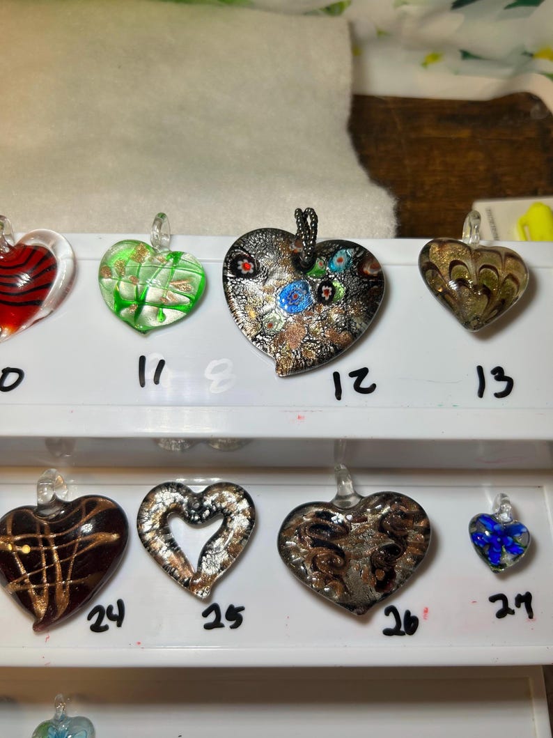 May include: A collection of heart-shaped glass pendants in various colors and designs. Some pendants have metallic accents, while others feature swirls, stripes, and floral patterns. Each pendant has a small loop for hanging and is numbered.