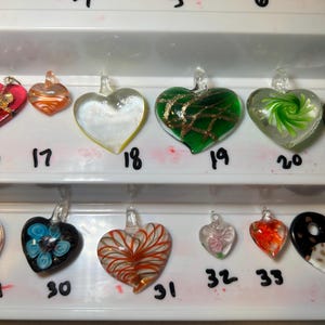 May include: A collection of heart-shaped glass pendants in various colors and designs. Some pendants are clear, others feature swirls, stripes, or floral patterns. Each pendant has a small loop for hanging. Numbers are written below each pendant.