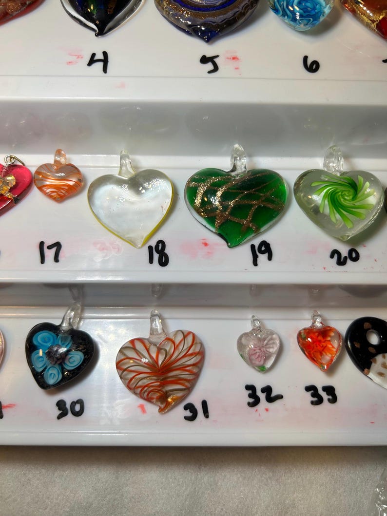 May include: A collection of heart-shaped glass pendants in various colors and designs. Some pendants are clear, others feature swirls, stripes, or floral patterns. Each pendant has a small loop for hanging. Numbers are written below each pendant.