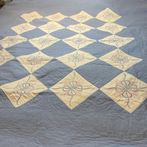 以下が含まれることがあります： 薄い青色のキルトで、ひし形の模様です。各ひし形は、ピンクと青色の刺繍の花柄模様のクリーム色の中心部を備えています。キルトはベッドの上に広げられています。