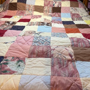 May include: A patchwork quilt with a variety of fabrics in shades of pink, cream, brown, and blue. The quilt is made of squares sewn together, creating a geometric pattern. The quilt is on a wooden bed frame.