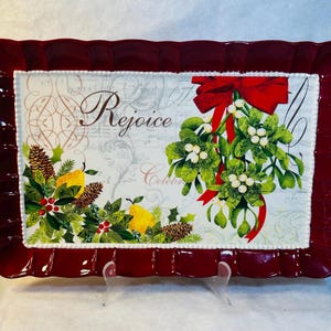 Puede incluir: Una bandeja rectangular roja con un borde blanco y un centro blanco con un diseño navideño con muérdago, peras y la palabra "Rejoice".