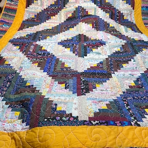 May include: A patchwork quilt with a diamond pattern in the centre. The quilt uses various fabrics in shades of blue, purple, and brown, with a yellow border. The quilt is handmade and appears to be made from fabric scraps.