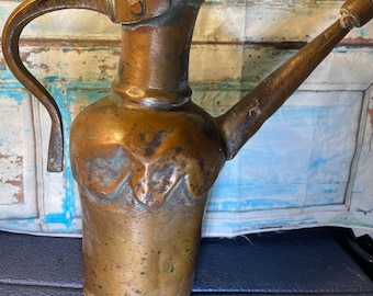 May include: A weathered copper pitcher featuring a long, slender spout, a curved handle, and a scalloped decorative band around the midsection. The metal surface displays a dark, aged patina with visible signs of oxidation and wear throughout.
