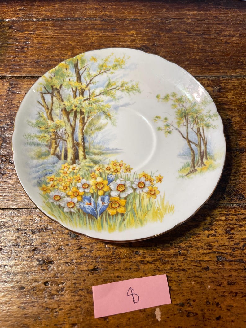 Może przedstawiać: Biały ceramiczny spodek z namalowaną sceną krajobrazową z ż&oacute;łtymi i białymi kwiatami, drzewami i błękitnym niebem. Spodek ma złoty brzeg i falisty brzeg. Og&oacute;lnym tematem są naczynia w stylu vintage.