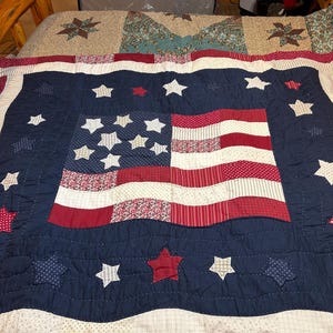 May include: A patchwork quilt featuring a central American flag design composed of red, white, and blue patterned fabrics. The flag is surrounded by a wide navy blue border adorned with multiple appliquéd stars in red, white, and blue fabric.