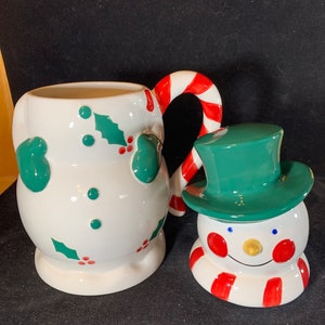 May include: A white ceramic snowman mug with a candy cane handle and holly leaf design. The mug is paired with a smaller snowman figurine with a green hat and red and white striped scarf.