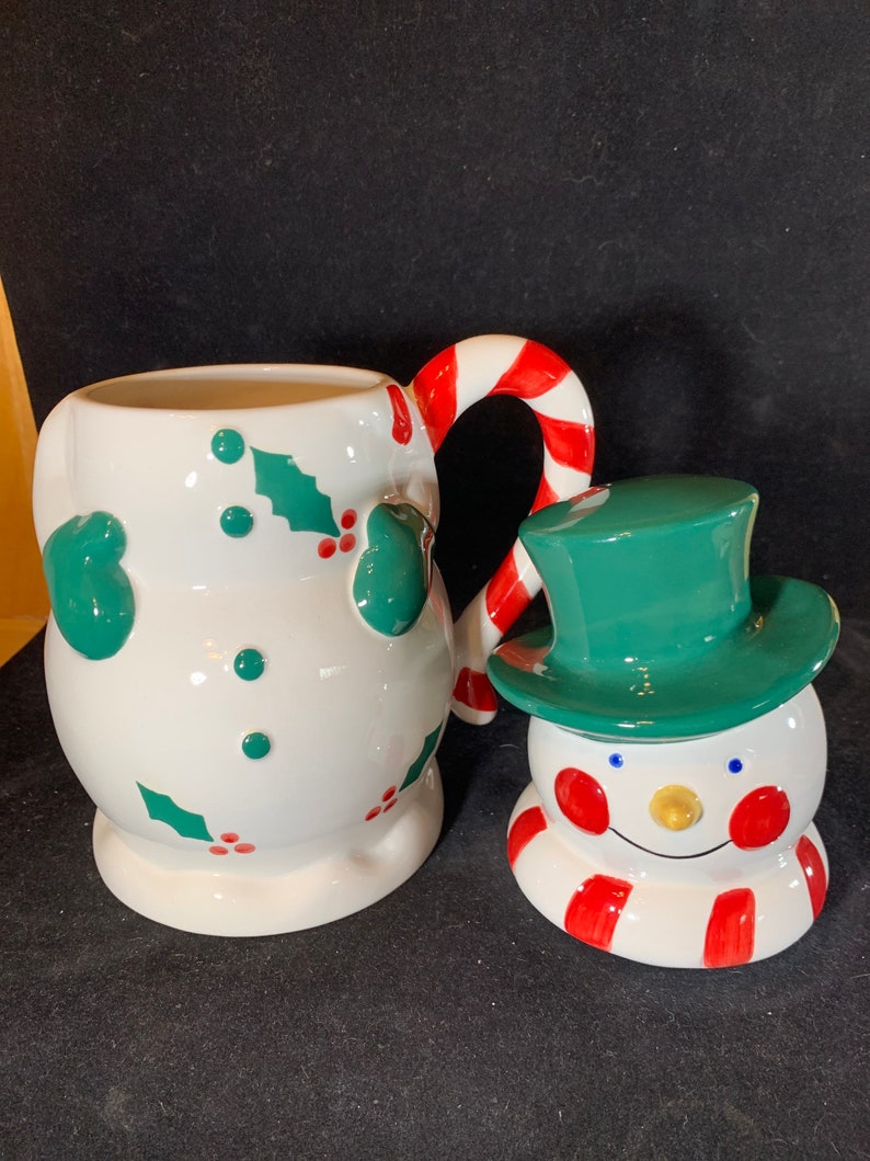 May include: A white ceramic snowman mug with a candy cane handle and holly leaf design. The mug is paired with a smaller snowman figurine with a green hat and red and white striped scarf.