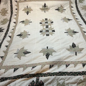 Può includere: Una trapunta fatta a mano con sfondo bianco e motivi a foglie nelle tonalità del verde, grigio e nero. La trapunta presenta un disegno centrale di quadrati ad incastro e un bordo con accenti floreali. La trapunta è su un telaio del letto in legno.