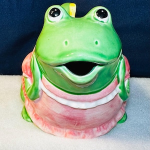 May include: A ceramic frog-shaped container with a wide open mouth. The frog is primarily green with black eyes and a pink and white dress-like design. The container has a lid and is likely a decorative item.