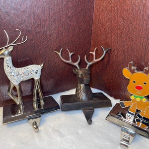 May include: Three Christmas stocking holders featuring deer designs. One is a silver deer with a decorative body, another is a bronze deer head, and the third is a cartoon reindeer with a red nose and green collar.