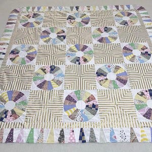 May include: A quilt featuring a repeating pattern of circular fan blocks and square blocks with tan and white stripes. The circular blocks are composed of multi-colored fabric wedges surrounding a white center. A border of small fabric triangles surrounds the quilt.