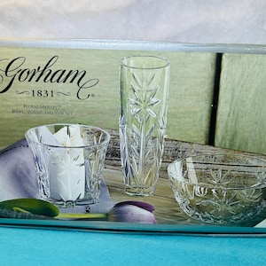 Può includere: Un set di tre pezzi in vetro trasparente con un motivo floreale. Il set comprende una ciotola, un portacandele e un vaso. La scatola è etichettata "Gorham 1831 Floral Majesty Bowl, Votive and Vase Set."