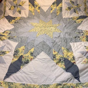 May include: A handmade quilt with a starburst design. The quilt features a central yellow star surrounded by gray, white, and blue patterned fabrics. Accents include floral and gingham patterns. The quilt is a cozy, decorative bedding item.