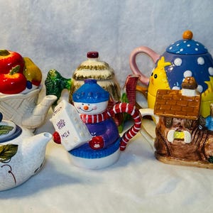 Puede incluir: Una colección de teteras decorativas de cerámica. Una tetera tiene forma de cesta de frutas, otra de muñeco de nieve con un cartón de leche, y otras presentan patrones y formas caprichosas. Las teteras son de diferentes tamaños.