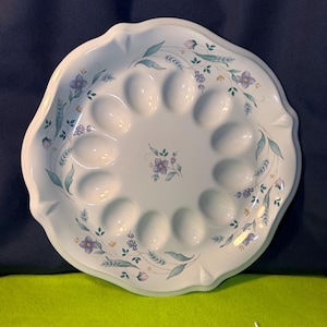 Puede incluir: Un plato de huevos de cerámica blanca con borde festoneado. El plato tiene 12 hendiduras para huevos y está decorado con un estampado floral en tonos morado, verde y amarillo. El plato es redondo y tiene un fondo azul claro.
