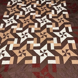 May include: A handmade quilt with a geometric pattern in shades of brown, tan, and white. The quilt features a repeating design of pinwheel and square shapes, creating a visually appealing and intricate pattern. The border is a darker shade of brown with red and white polka dot accents.