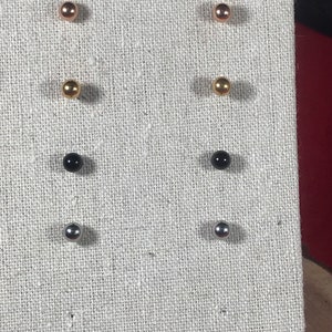 May include: Six pairs of stud earrings displayed on a fabric-covered board. The earrings are in four different colors: gold, rose gold, black, and silver.
