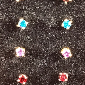 May include: A set of eight small, colorful gemstone earrings. The earrings are set in gold and feature a variety of colors, including red, blue, purple, and black.