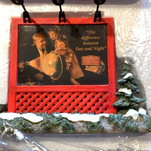 May include: A red and white miniature billboard with a vintage advertisement for Eveready Flashlights and Batteries. The ad features a family looking at a globe and the text "The difference between Day and Night".