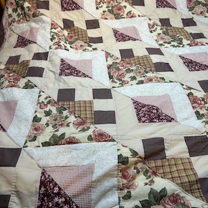 May include: A patchwork quilt with a floral and geometric pattern. The quilt features squares and triangles in shades of beige, brown, and burgundy. Some squares have a floral print, while others have a plaid or solid design. The quilt is likely made of cotton.