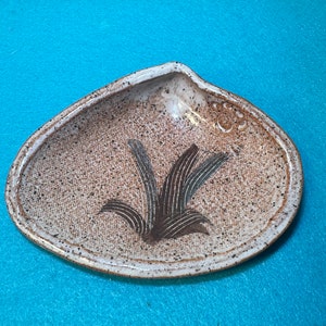 May include: A white ceramic dish shaped like a seashell with a brown and green floral design. The dish has a speckled glaze and a slightly raised rim.