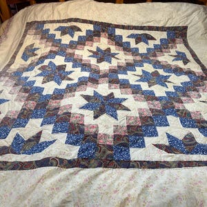 以下が含まれることがあります： 手作りのキルト、星柄。キルトは、クリーム色の背景に青、赤、ピンクの星と正方形のデザインが特徴です。ボーダーは濃い青のペイズリープリントです。キルトは、花柄のシーツが敷かれたベッドの上にあります。