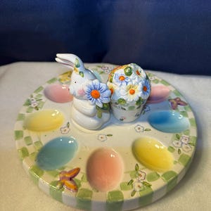 以下が含まれることがあります： ウサギと花の模様が施された陶器製イースターエッグホルダー。エッグホルダーは緑と白のチェック柄の縁取りで、パステルカラーの卵がいくつか入っています。ホルダーには蝶も描かれています。