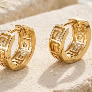 May include: A pair of gold-toned hoop earrings featuring a cut-out Greek key pattern along the outer surface. The earrings have a hinged closure mechanism and a polished metallic finish, showcasing a repeating geometric design around the circular band.