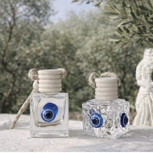May include: Two clear glass car air freshener bottles with wooden caps and hanging ropes. The left bottle is smooth and square, while the right bottle has a textured, faceted surface. Both feature a blue, white, and black evil eye bead attached.