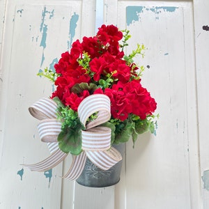 May include: A red geranium plant in a metal pot with a brown and white striped ribbon bow. The plant is hanging on a white door with blue paint chipping.