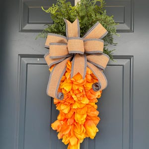Pode incluir: Uma coroa de porta de Páscoa em forma de cenoura com pétalos de tecido laranja, um laço de juta com fita xadrez preta e branca e folhagem verde.