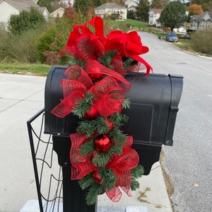Pode incluir: Uma caixa de correio preta decorada com um laço vermelho e uma guirlanda para a época natalina. A guirlanda é feita de malha vermelha, ramos de pinheiro verdes e enfeites vermelhos.