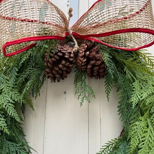 Pode incluir: Uma grinalda verde com agulhas de pinho e pinhas marrons decoradas com um la&ccedil;o de juta vermelho e marrom.