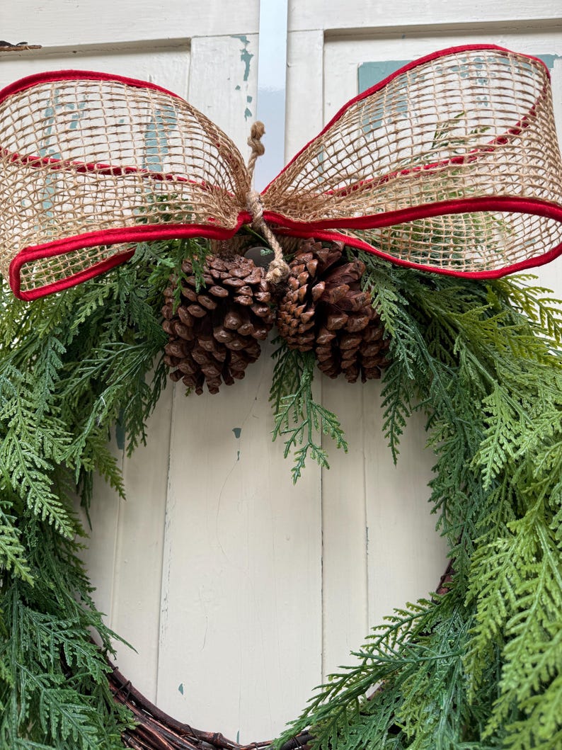 Pode incluir: Uma grinalda verde com agulhas de pinho e pinhas marrons decoradas com um la&ccedil;o de juta vermelho e marrom.