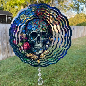May include: A colorful metal wind spinner with a skull design surrounded by flowers. The spinner has multiple layers and is suspended from a chain with crystal beads.