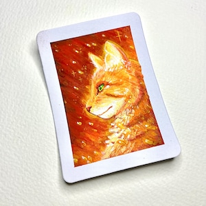 May include: A rectangular magnet featuring a vibrant painting of an orange tabby cat in profile. The cat has bright green eyes and is set against a fiery red and orange background with small, glowing yellow highlights and cross-shaped sparkles.