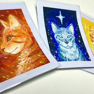 May include: Three rectangular art prints in clear protective sleeves. The left print shows an orange cat against a red background. The centre print features a blue cat under a white star on a dark blue background. The right print depicts a light orange cat on a yellow background.