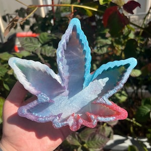 Puede incluir: Una bandeja con forma de hoja de cannabis en tonos azules, morados y rojos. La bandeja de resina tiene un acabado brillante y se sostiene en una mano. La bandeja está diseñada con un efecto en capas, creando profundidad y dimensión. El fondo presenta follaje verde.