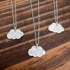 Puede incluir: Tres collares con colgantes en forma de nube y cadenas plateadas. Las nubes son blancas con efecto marmoleado. Los collares se exhiben sobre una superficie de madera oscura. Las cadenas son delicadas.