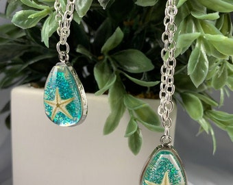 May include: Two silver necklaces with teardrop pendants. Each pendant features a starfish encased in clear resin with teal glitter. The pendants hang from a silver chain, creating a nautical theme.