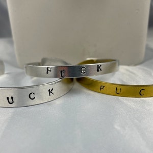 May include: Three metal cuff bracelets in silver and gold tones. Each bracelet has the word "FUCK" stamped across the front. The bracelets are open and adjustable.