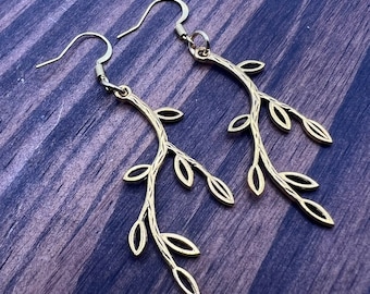 May include: A pair of gold-colored leaf branch earrings. Each earring features a curved branch design with multiple small, delicate leaves. The earrings have a hook closure and are displayed against a dark wood grain background.