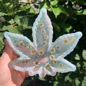 May include: A leaf-shaped resin tray with a shimmering, iridescent finish. The tray is filled with small, dried daisies, creating a unique decorative piece. The edges have a light blue and white color.