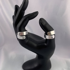 Puede incluir: Dos anillos ajustables de color plateado, cada uno con las letras "F" y "U" estampadas. Los anillos se exhiben en un soporte para joyas negro con forma de mano sobre un fondo blanco.