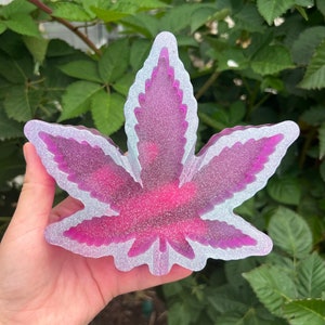 May include: A decorative ashtray in the shape of a cannabis leaf. The ashtray is a vibrant pink with glitter accents and a white outline. The leaf shape is detailed and the ashtray is held in a hand against a backdrop of green foliage.