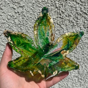 Puede incluir: Una hoja de resina verde y dorada con un diseño marmoleado. La hoja tiene forma de hoja de marihuana y tiene detalles dorados.