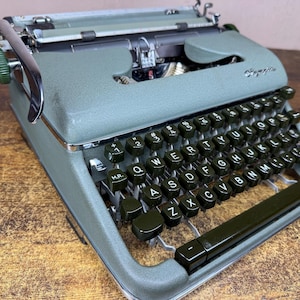 Peut inclure: Une machine à écrire portable Olympia vert sauge dotée d'un clavier QWERTY avec des touches circulaires vert foncé, un levier de retour de chariot chromé et un bouton vert sur le côté gauche. Le boîtier métallique a une finition texturée.
