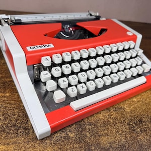 May include: A portable Olympia typewriter featuring a vibrant red metal body with white accents. The device includes a QWERTY keyboard with white keys, black lettering, and a prominent Olympia brand label on the top left side of the frame.